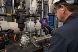 Technician using a thermal camera to inspect a steam trap and valve station for maintenance or replacement needs.